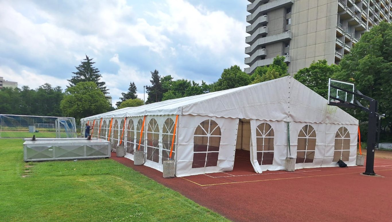 Veranstaltungszelt auf Sportplatz Zelt auf einem Sportplatz, umgeben von Bäumen und einem Wohngebäude im Hintergrund.