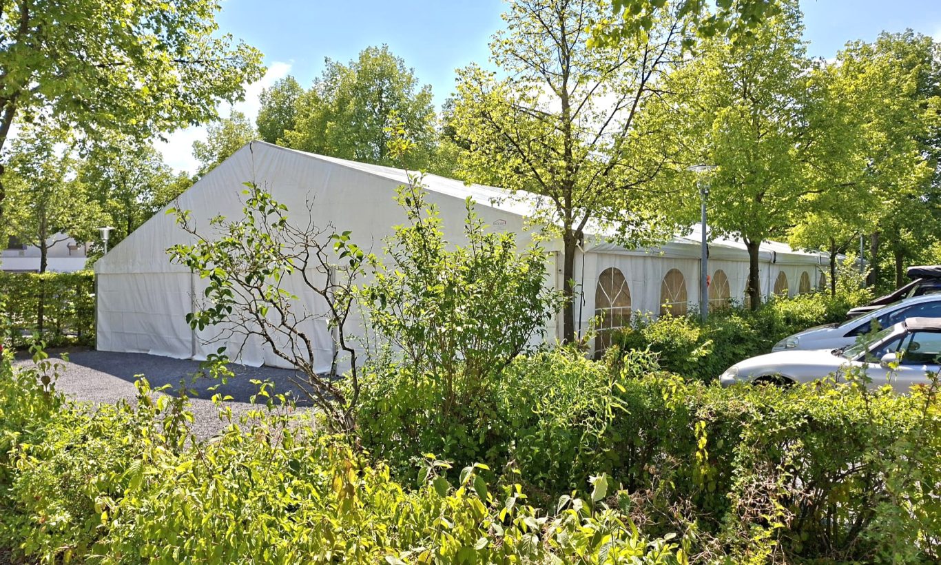 Großzelt im Grünen Weißes Großzelt inmitten von Bäumen und Büschen, mit parkenden Autos in der Nähe.