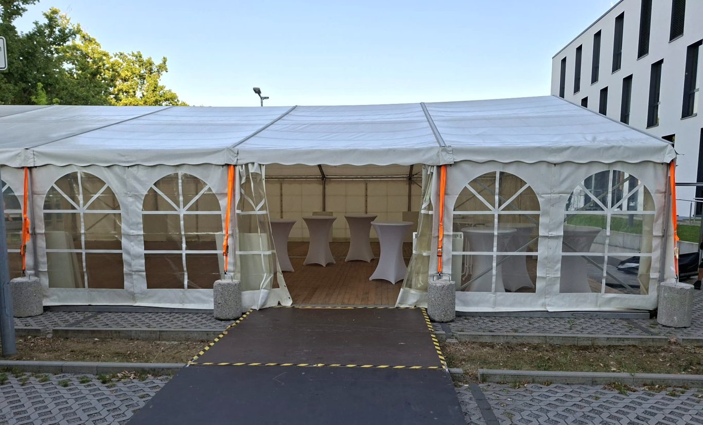 6m breites Zelt mit Stehtischen Ein Zelt mit Stehtischen, umgeben von Gräsern und modernen Gebäuden im Hintergrund.