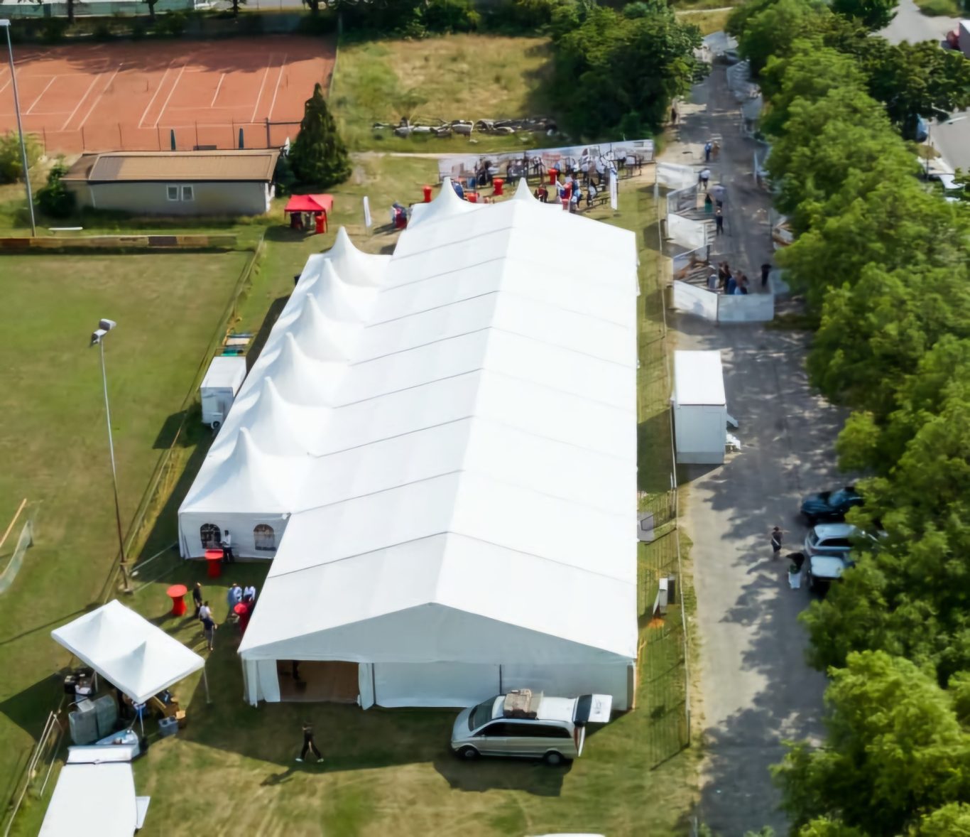 Großzelt mit Pagoden auf Sportplatz Großes, weißes Zelt auf einem Sportplatz, umgeben von Bäumen und Parkplätzen.