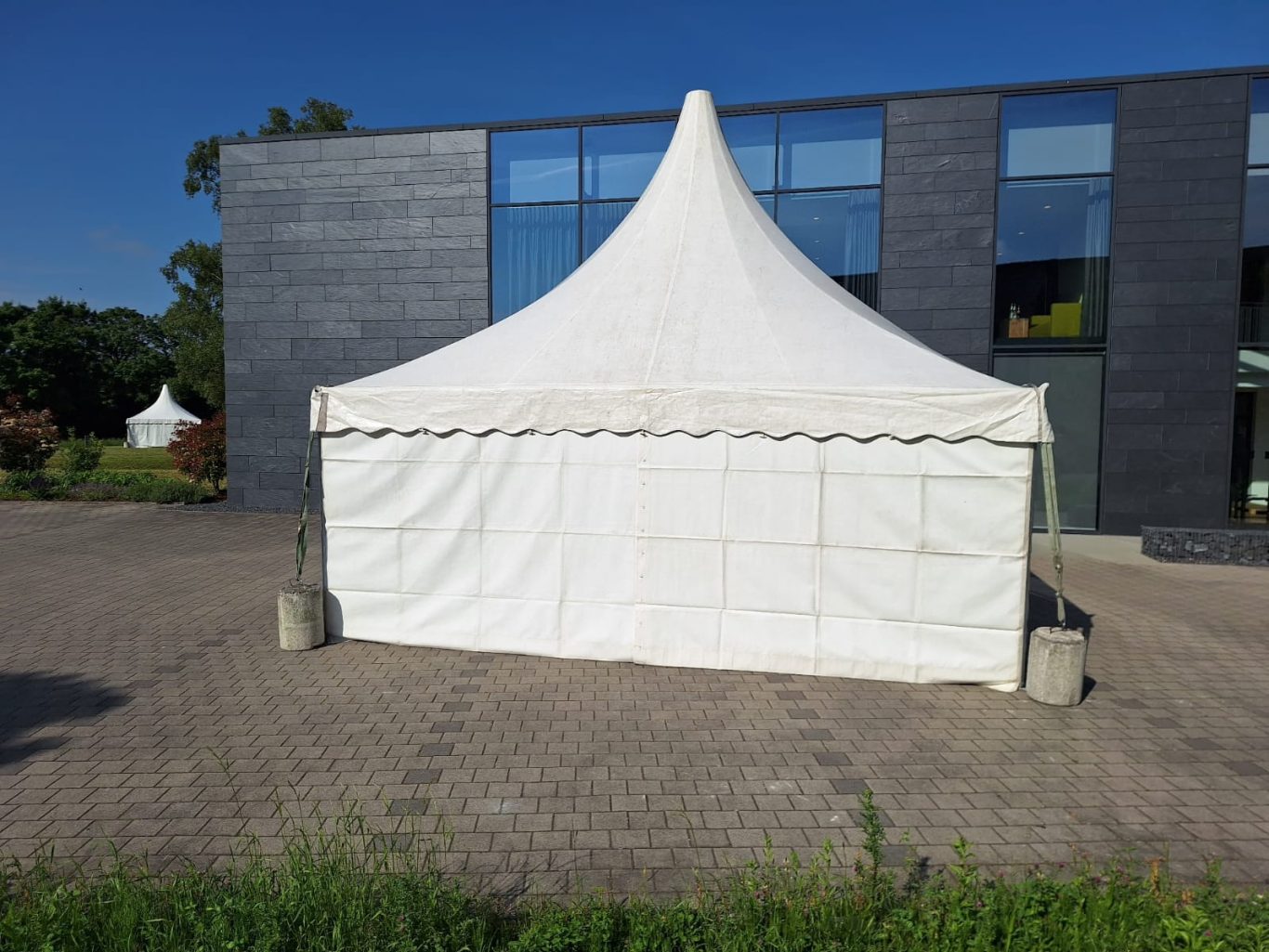 Großes Pagodenzelt vor Firmen-Eingang Weißes Zelt mit spitzem Dach auf gepflasterter Fläche vor einem modernen Gebäude.