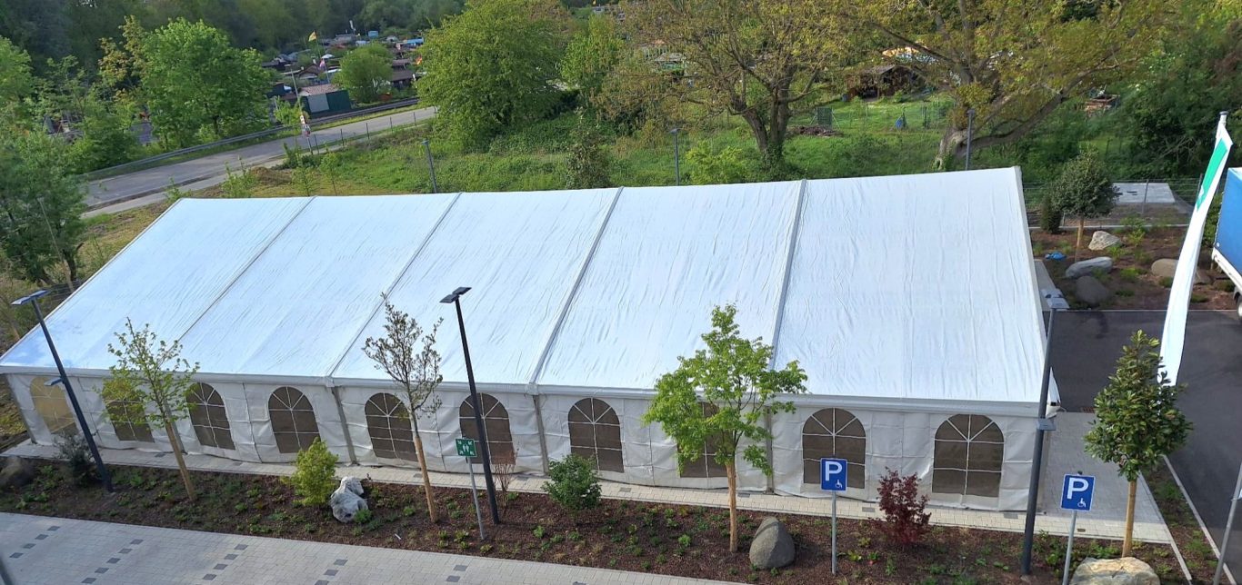 Großzelt für Firmenveranstaltung Großes Zelt mit weißem Dach, umgeben von Bäumen und einer Straße im Hintergrund.