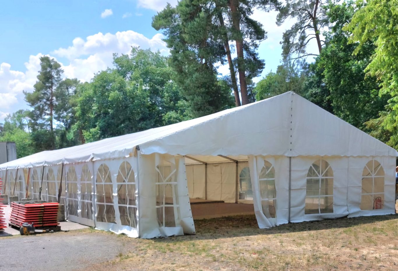 Partyzelt mit Fensterplanen Großes, weißes Zelt mit durchsichtigen Seitenwänden, umgeben von Bäumen.