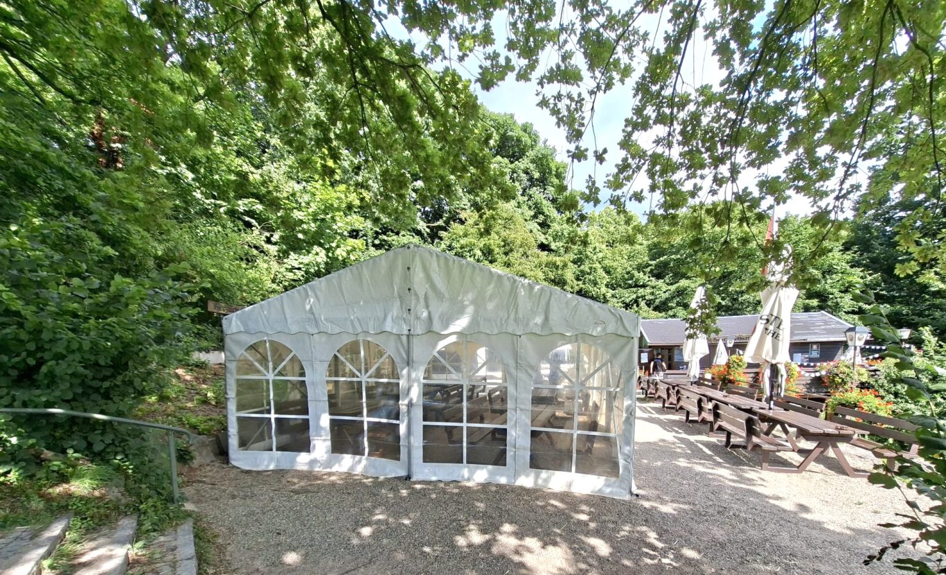 Zelt vor Restaurant im Grünen Weißes Festzelt umgeben von Bäumen in einer Außenanlage.