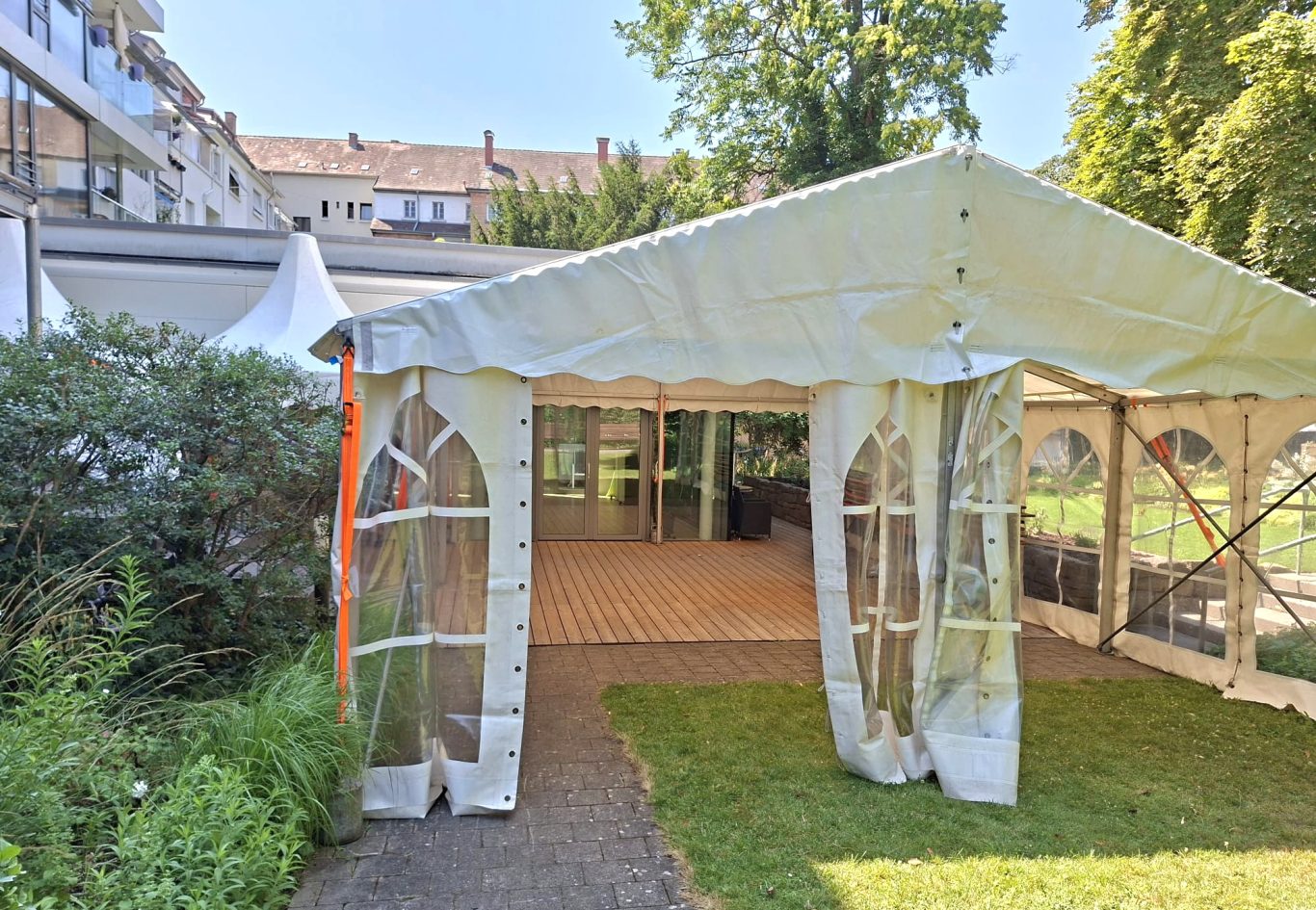 Zelt als Eingangsbereich Zelt mit offenen Seiten in einem Garten, umgeben von Grün und Gebäuden im Hintergrund.
