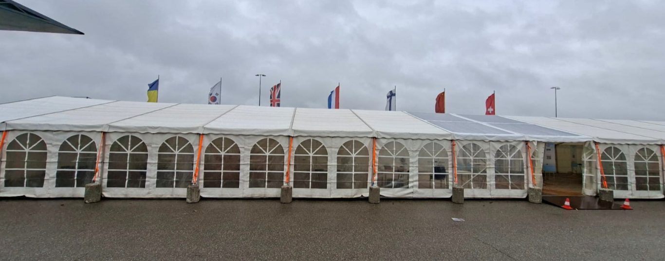 10m x 39m Zelt mit transparentem Dach Großes Zelt mit mehreren Fenstern, Fahnen und grauem Himmel im Hintergrund.
