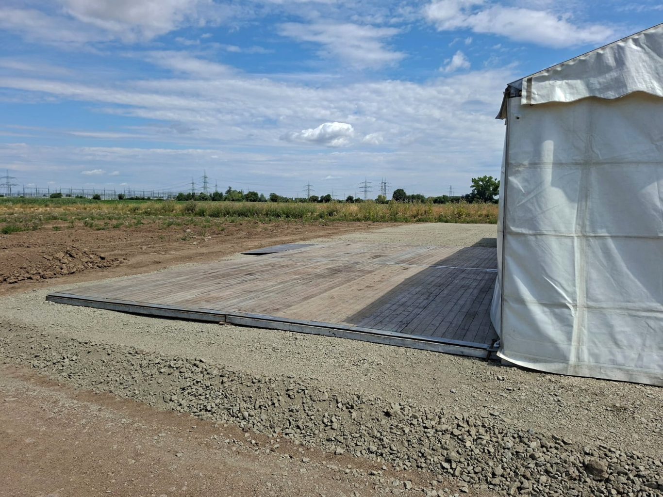 10m Zelt und Holzterrasse Baufeld mit Planenunterlage und Zelt in ländlicher Umgebung unter blauem Himmel.