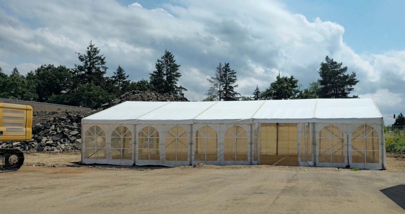 Zelt für Bergfest auf Baustelle Ein großes Zelt auf einer befestigten Fläche, umgeben von Bäumen und Wolken.