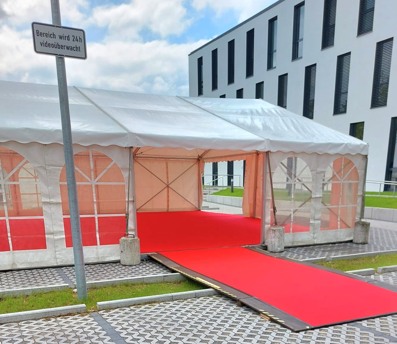 Kleines Empfangszelt mit Stehtischen Zelt mit rotem Teppich vor modernem Gebäude, ausgelegt für eine Veranstaltung.